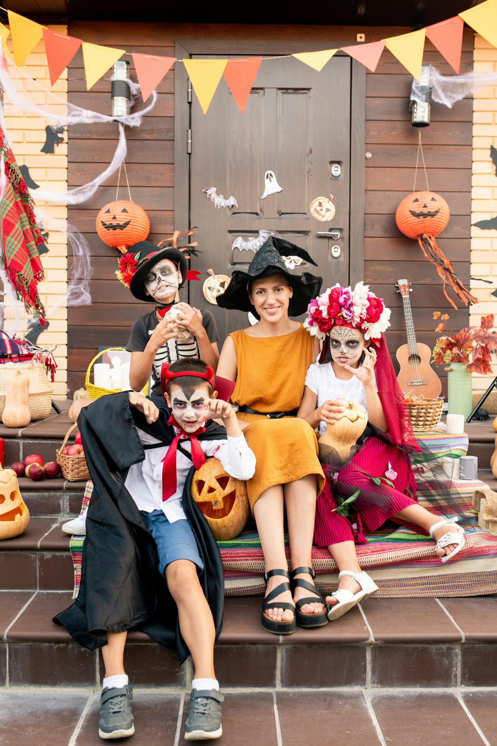 A mother and three children in Halloween costumes and face paint on a porch decorated with paper lanterns, bunting, and cobwebs